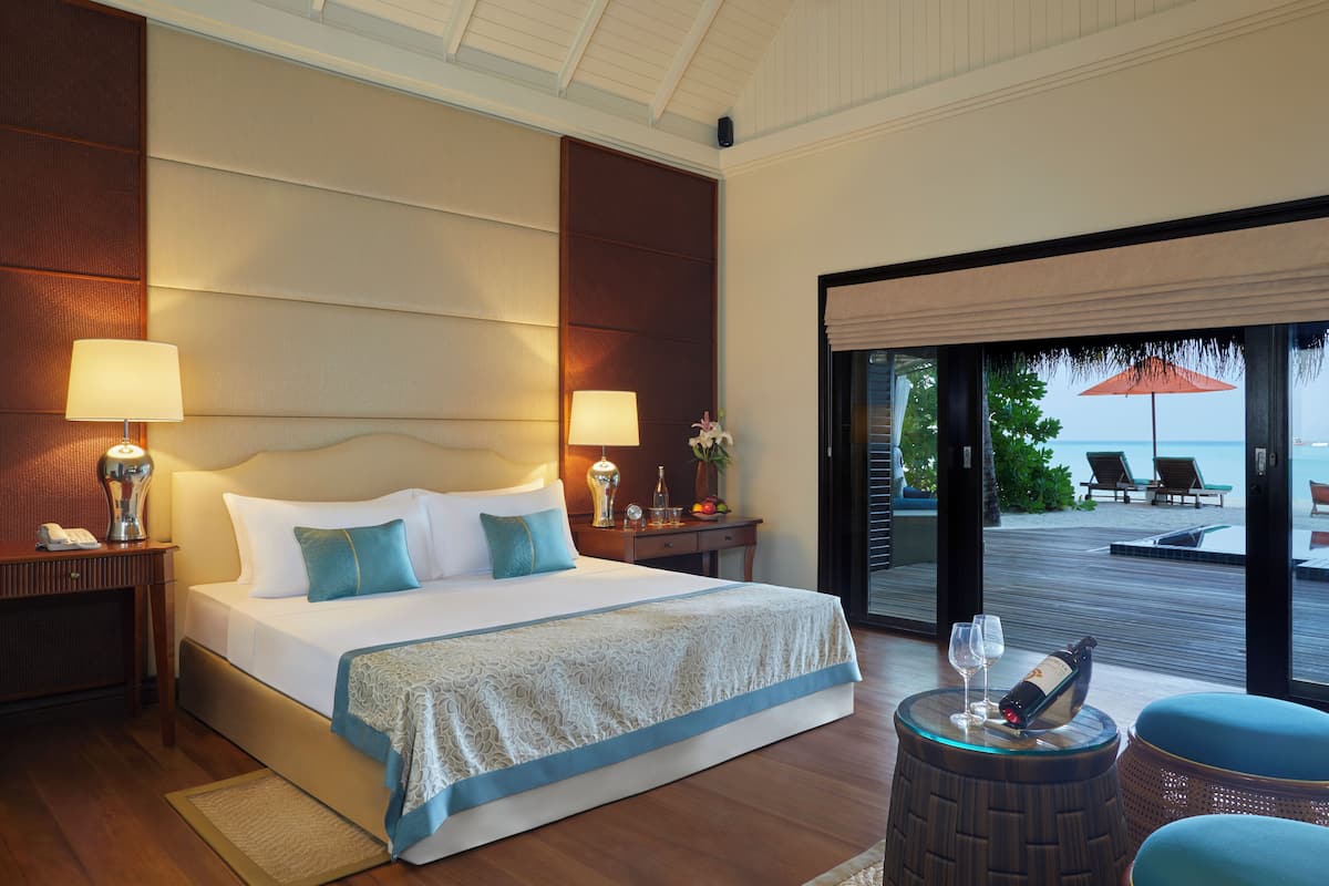 Ein Hotelzimmer mit einem großen Bett, zwei Nachttischlampen, einem kleinen Tisch mit Flasche und Gläsern sowie Blick auf den Strand durch eine Schiebetür.