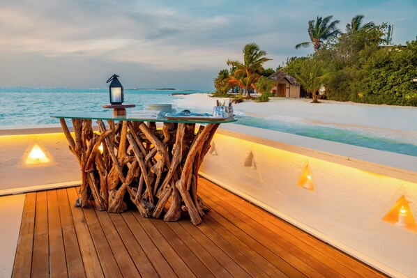Terraza de madera con una mesa de vidrio y un farol, con vistas a una playa con palmeras y cielo despejado.