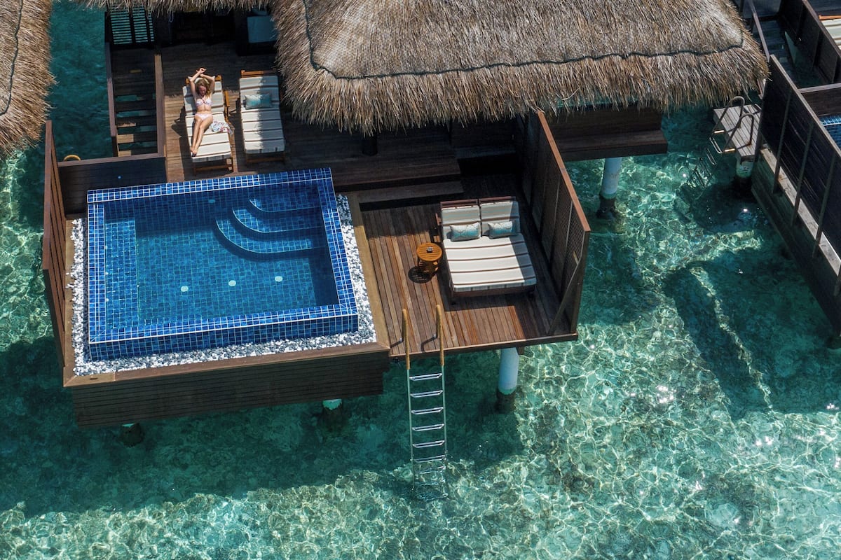 Bungalows auf Stelzen mit Reetdächern, einem Swimmingpool und Gartenmöbeln aus Holz.