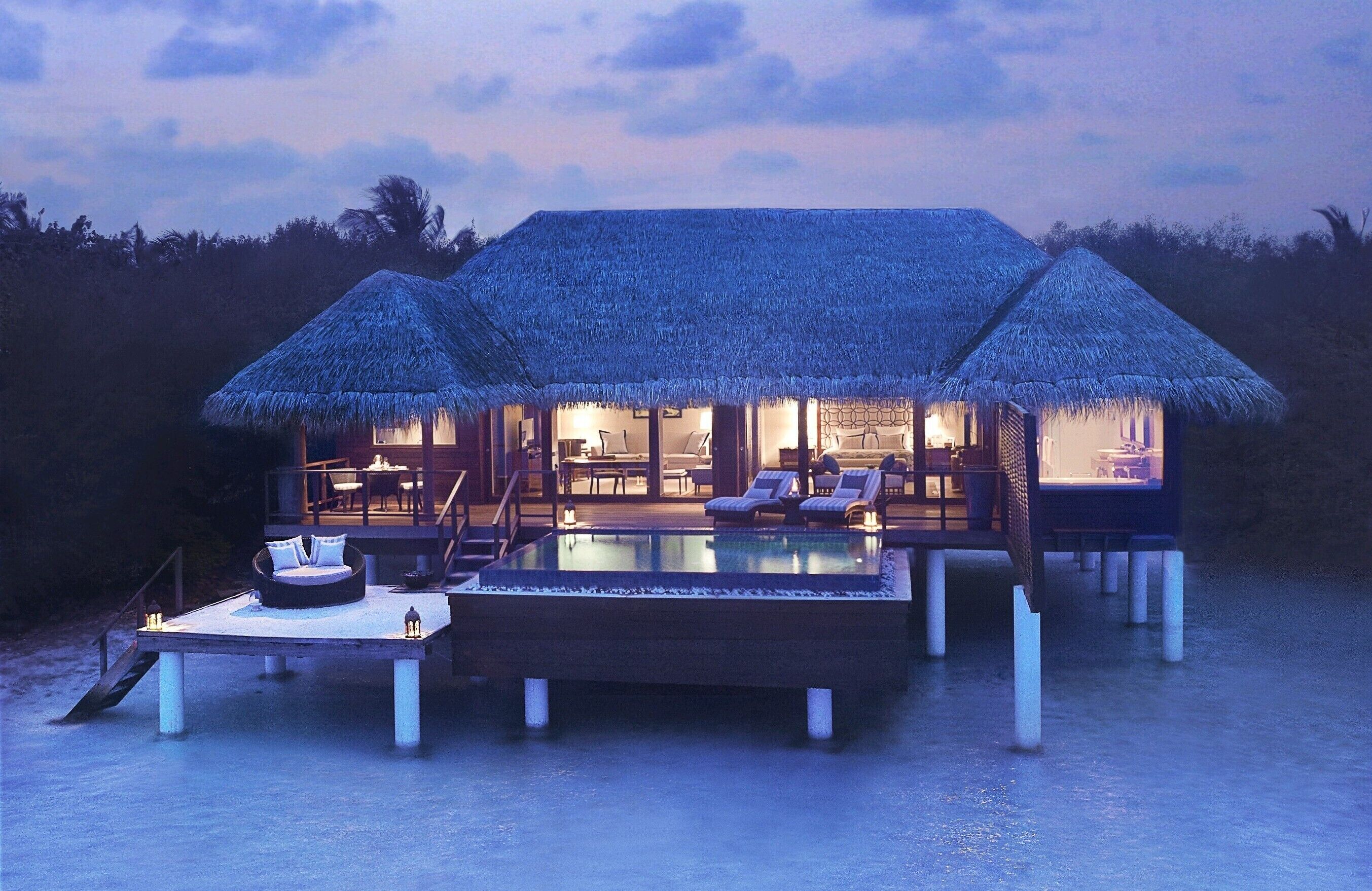 A beachfront overwater bungalow with a thatched roof, an infinity pool, and a dining area.