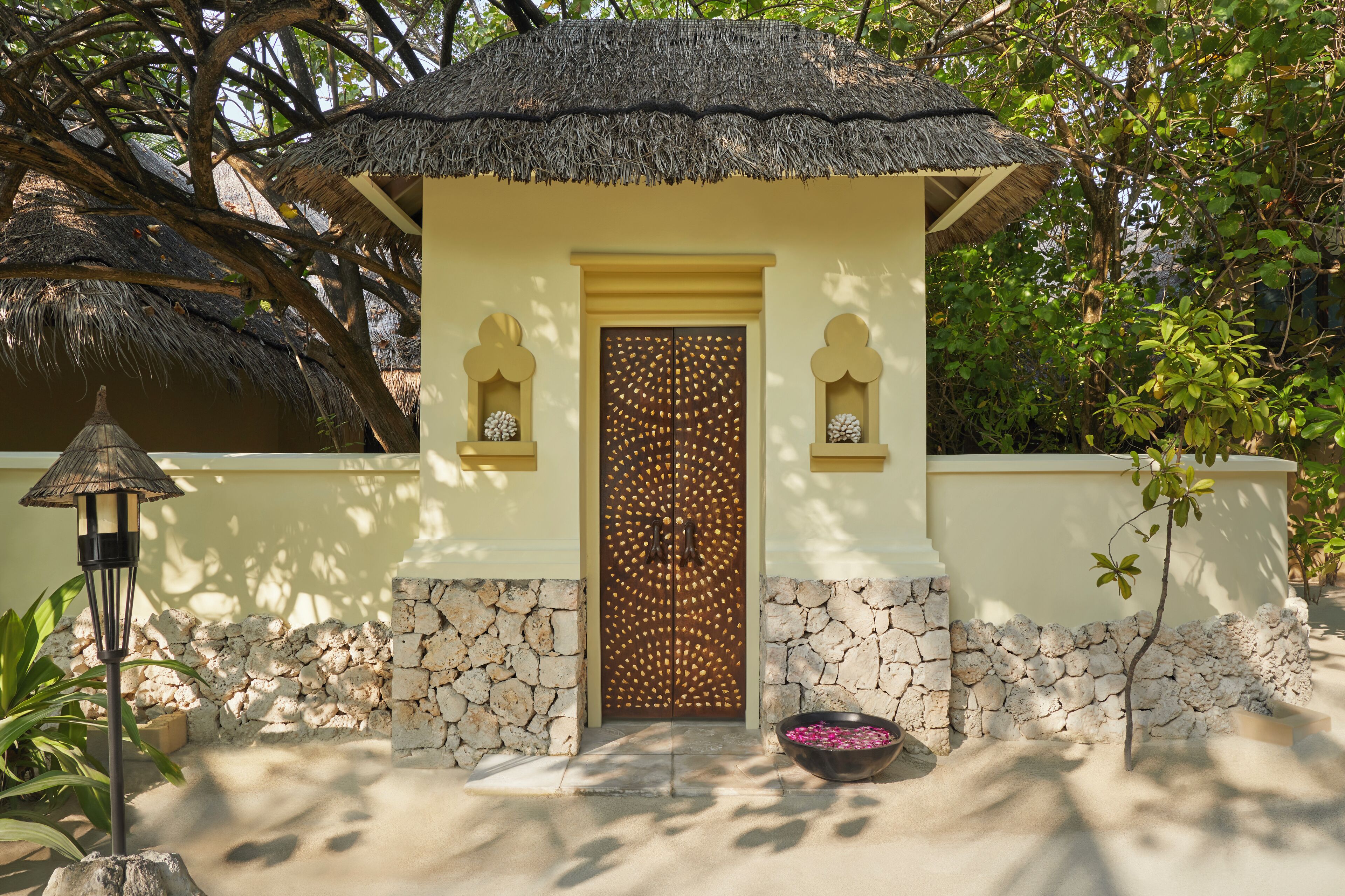 A small, thatched-roof structure with a stone wall and a decorative door, surrounded by greenery.