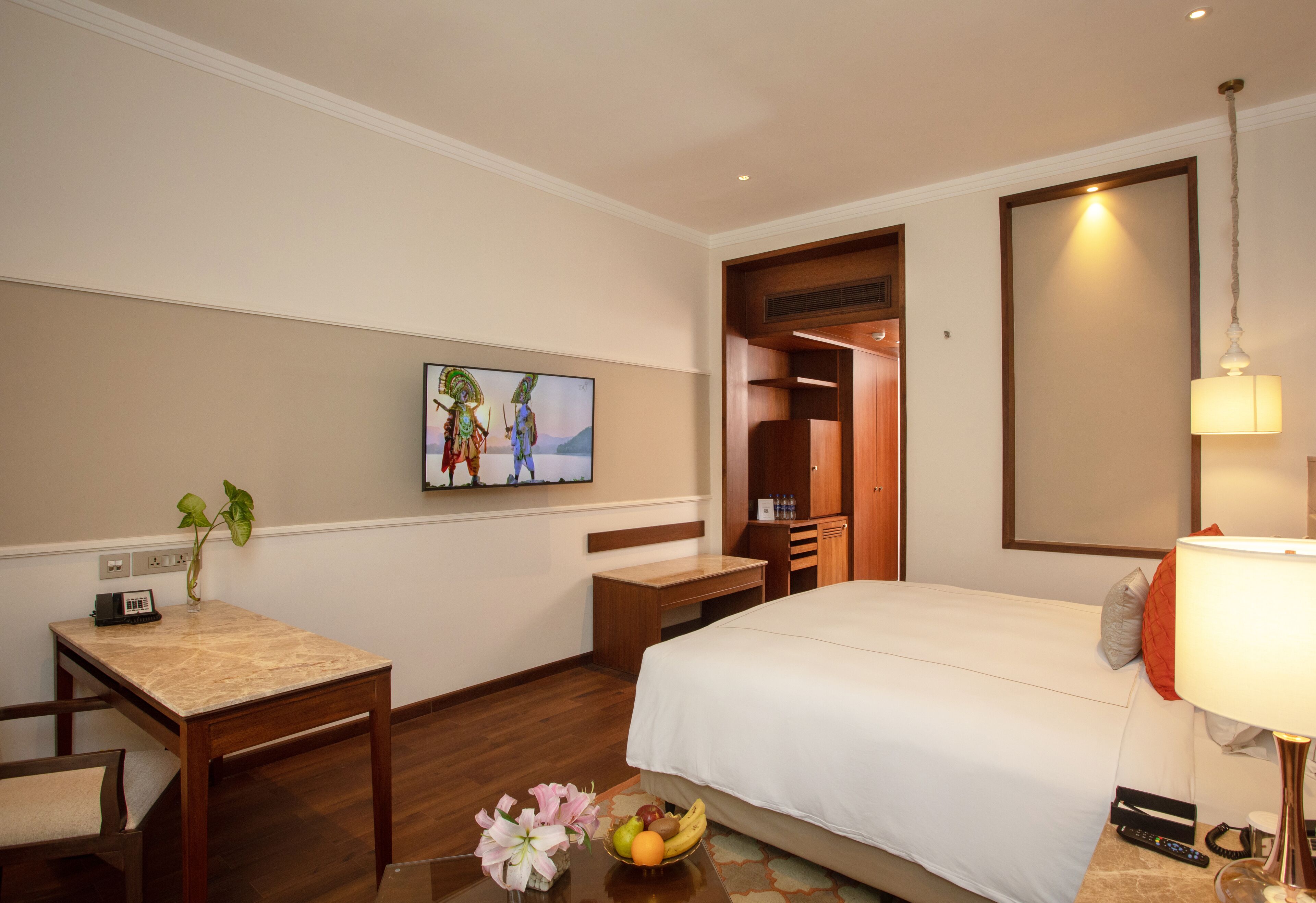 A hotel room with a bed, a desk, a chair, a small table with a vase of flowers, and a painting on the wall.