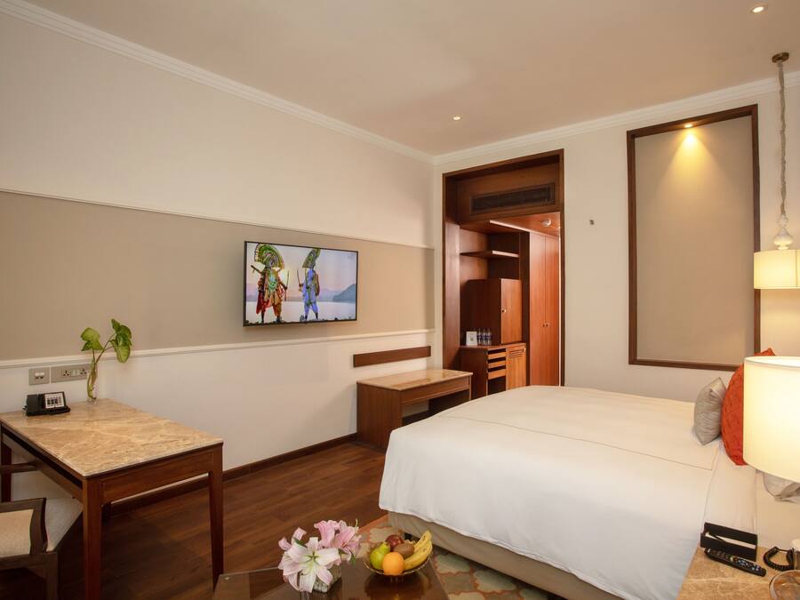 Una habitación de hotel con cama, escritorio, silla, una pequeña mesa con un florero y un cuadro en la pared.