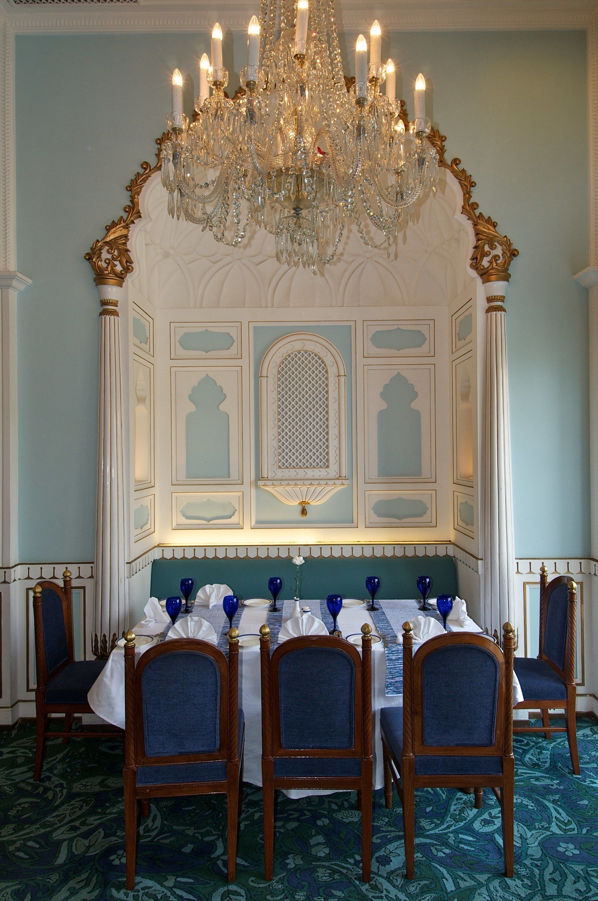 Comedor con candelabro, paneles de pared ornamentados y una mesa puesta para seis personas con sillas azules.