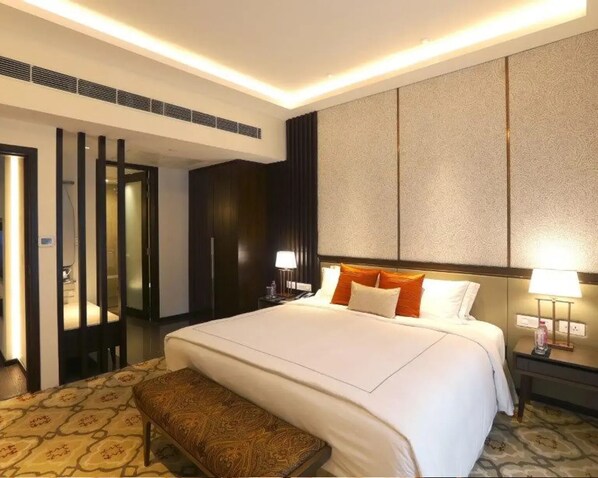 Una habitación de hotel con una cama grande, una banca, una mesita de noche con lámpara y una pared con textura.