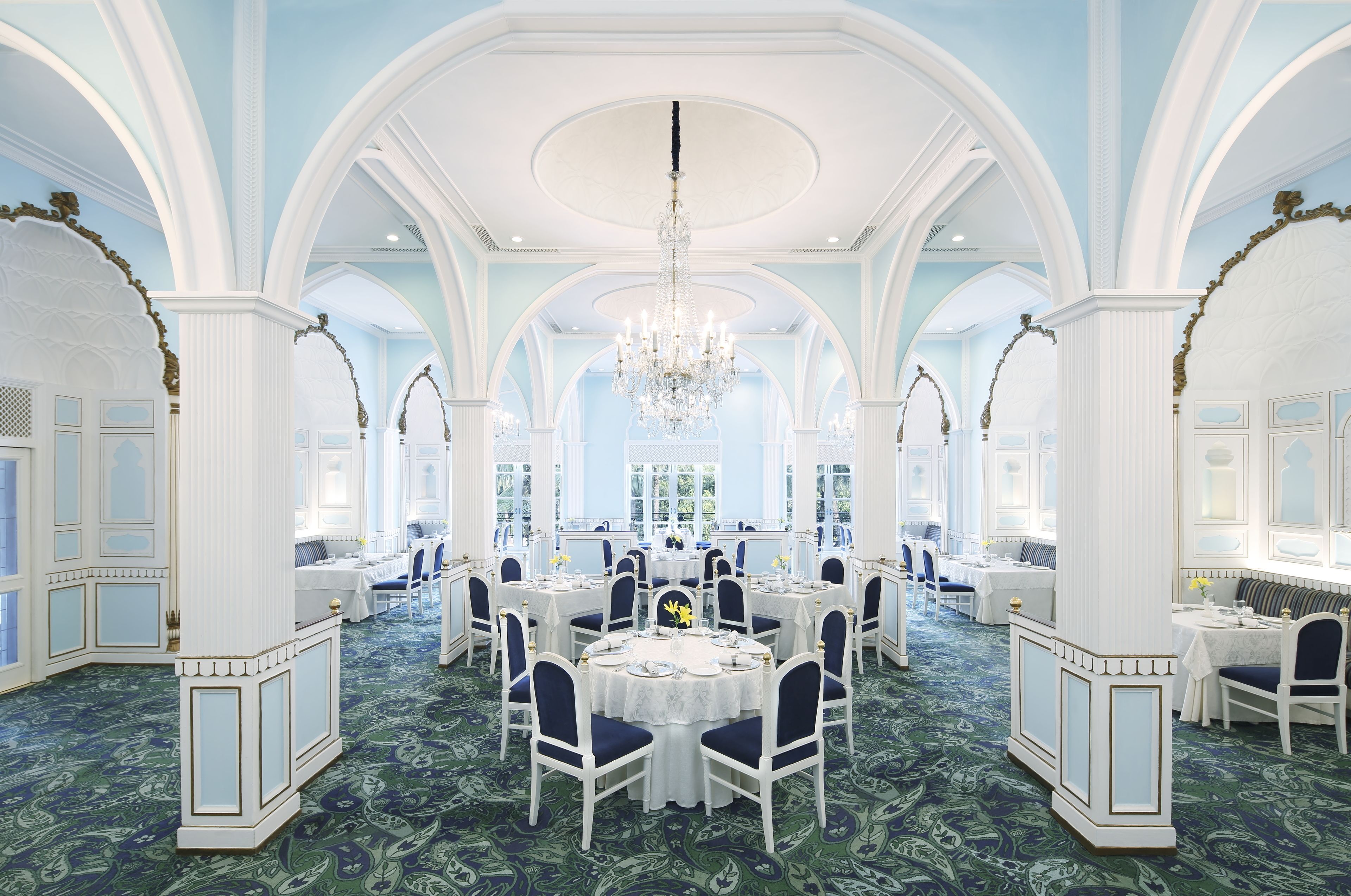 Un gran salón comedor con techos altos, candelabros y columnas ornamentales. Las mesas están dispuestas para una comida formal.