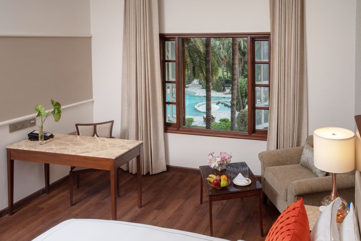 A hotel room with a wooden dining table, a sofa, a small table with fruit and flowers, and a view of a swimming pool through a window.