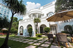 A white building with arched windows and columns, surrounded by a well-maintained garden with a paved walkway and outdoor seating area.