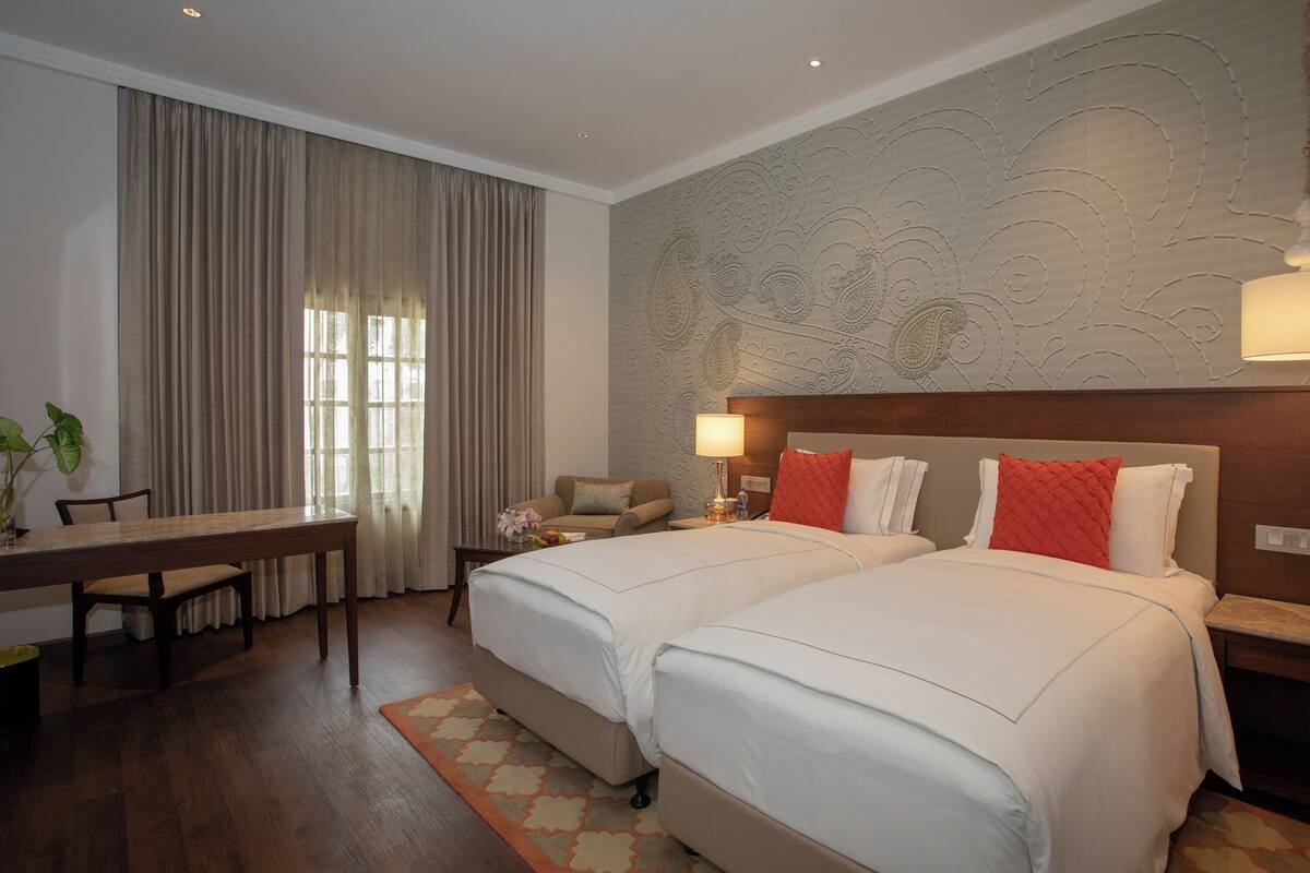 A hotel room with two beds, a dining table, a sofa, and a large window with curtains.