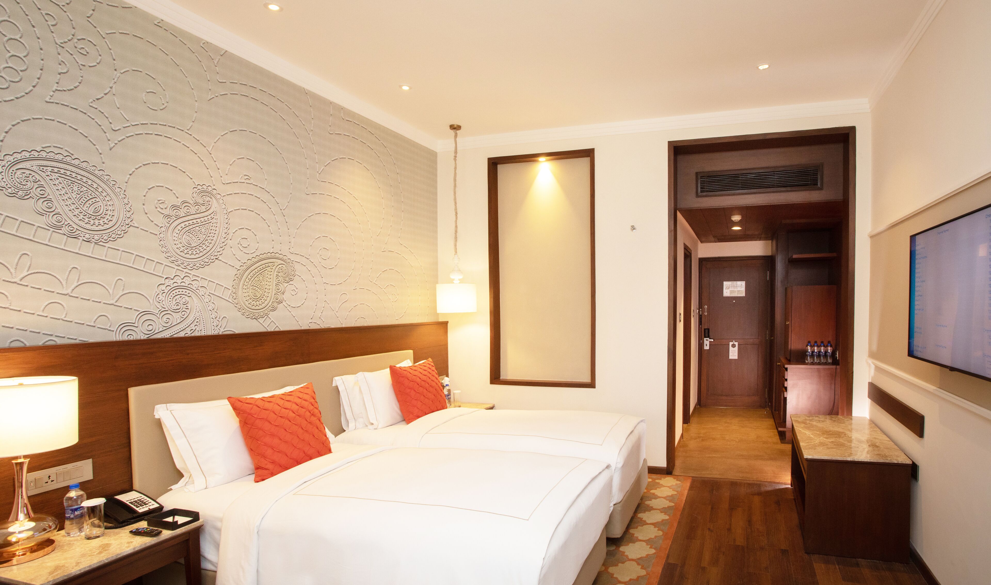 Een hotelkamer met twee bedden, een nachtkastje met een lamp, een muur met ingewikkelde patronen, een grote spiegel en een televisie aan de muur.