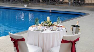 A poolside dining setup with white tables and chairs, red sashes, and a champagne bucket.