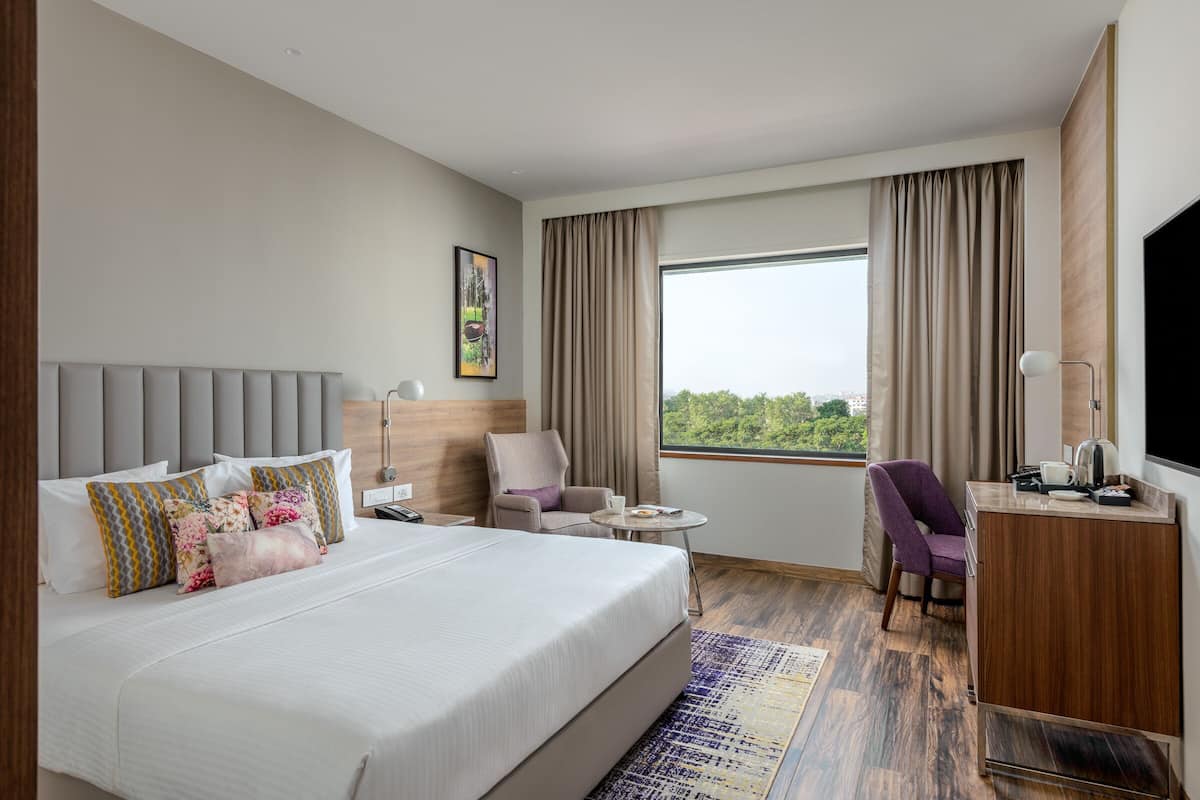 A hotel room with a bed, two chairs, a desk, a TV, and a window with curtains.