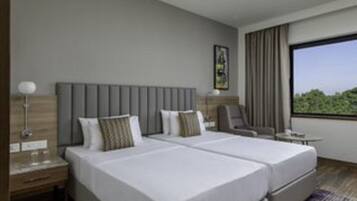A hotel room with two beds, a nightstand, a chair, a small table, and a large window offering a view of greenery.