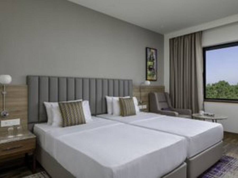 A hotel room with two beds, a nightstand, a chair, a small table, and a large window offering a view of greenery.