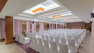 Un salón de conferencias con hileras de sillas blancas, una alfombra estampada e iluminación empotrada en un techo con tragaluces.
