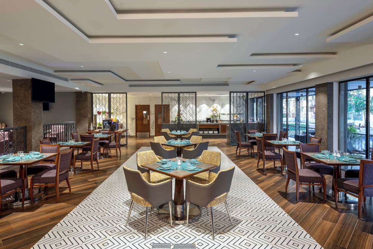 A spacious restaurant with multiple dining tables and chairs, a patterned floor, and large windows offering a view of the outdoors.