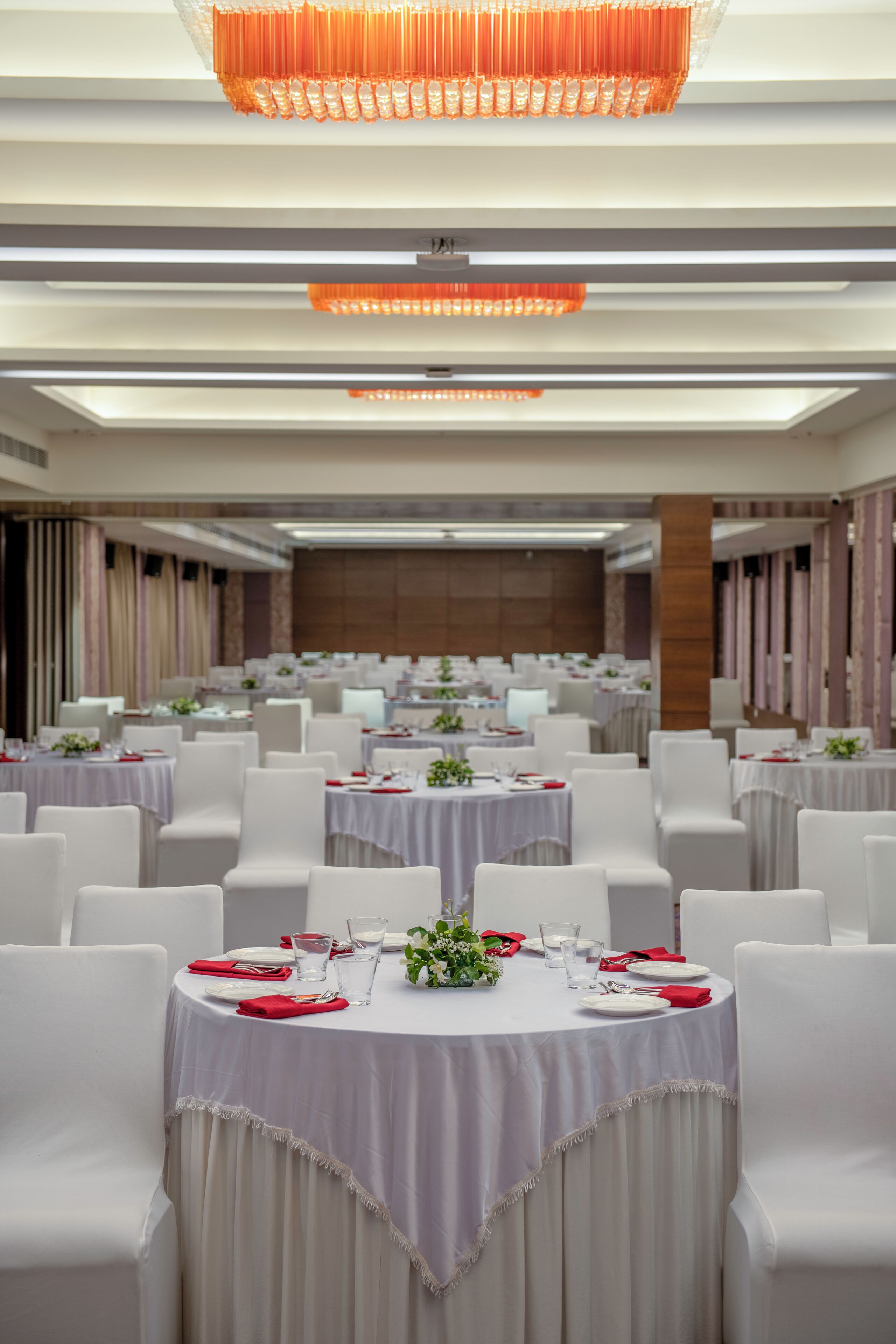 Uma sala de banquetes com mesas redondas, cadeiras brancas e guardanapos vermelhos.
