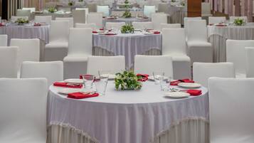 Uma sala de banquetes com mesas redondas, cadeiras brancas e guardanapos vermelhos.