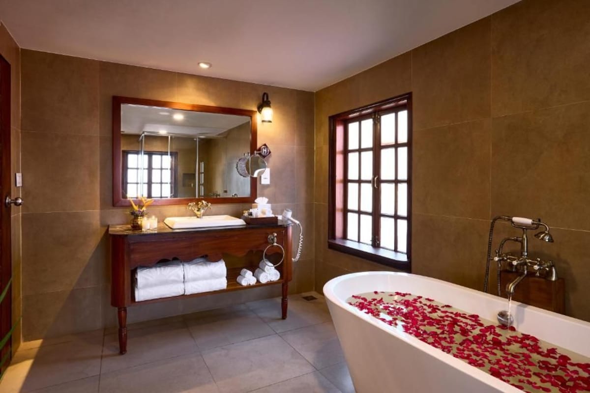 Une salle de bain avec une baignoire îlot remplie de pétales de roses, un meuble-lavabo en bois avec un miroir et une fenêtre donnant sur un paysage agréable.