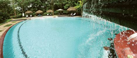 A large outdoor swimming pool with a waterfall feature, surrounded by lounge chairs and thatched umbrellas.