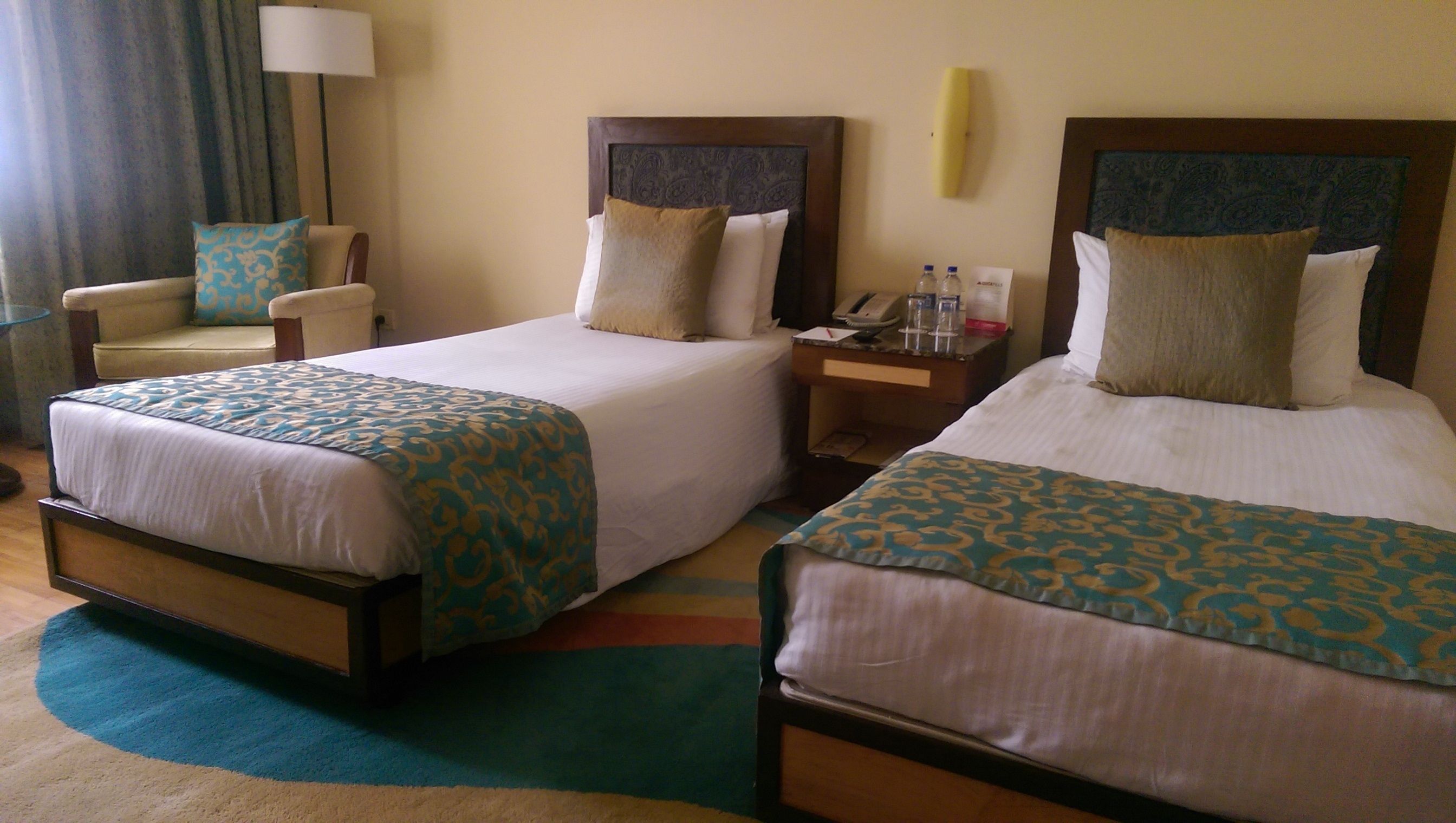 Habitación de hotel con dos camas individuales, una silla, una mesita de noche y una lámpara.