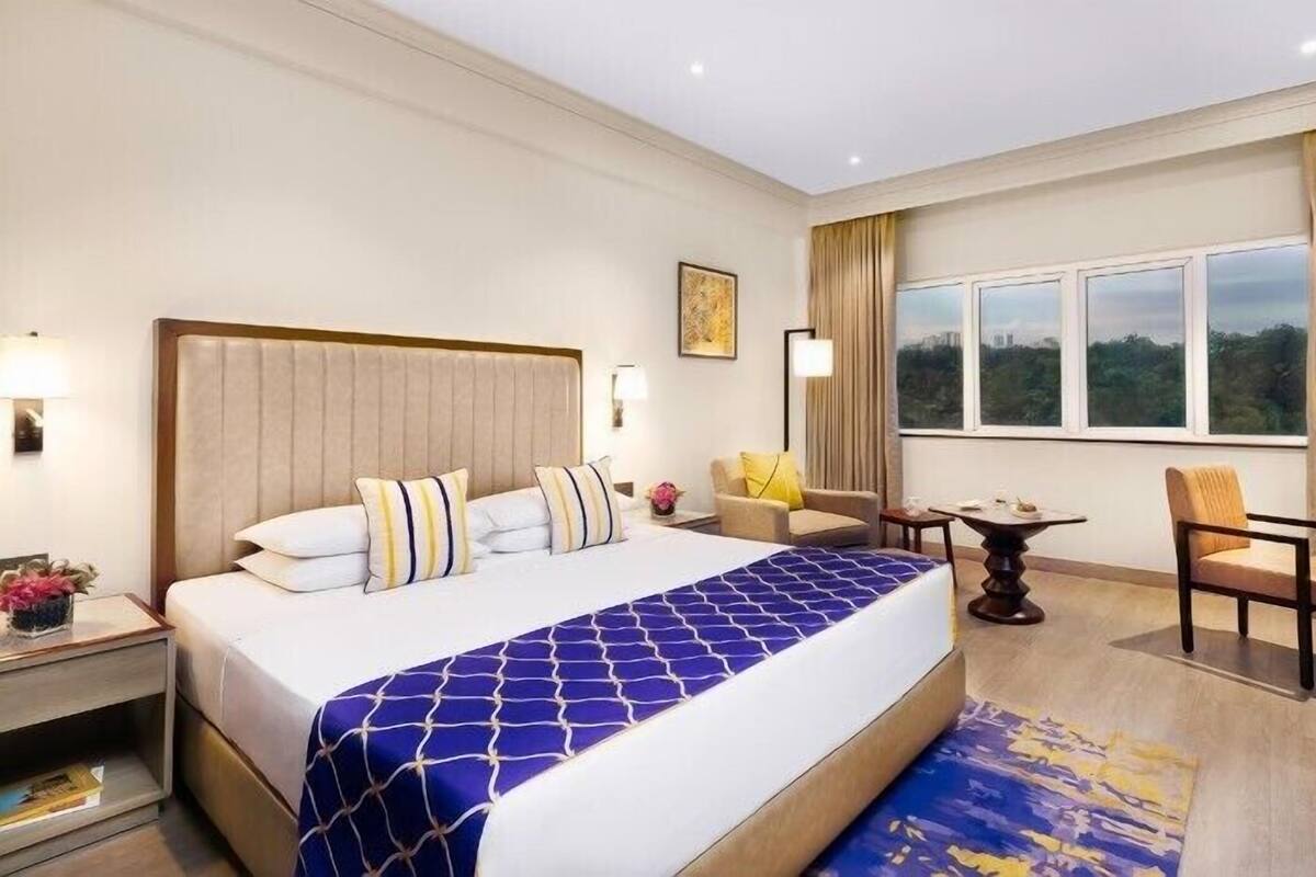 Habitación de hotel con una cama grande, una mesita, dos sillas, una mesita de noche y una ventana con vistas.