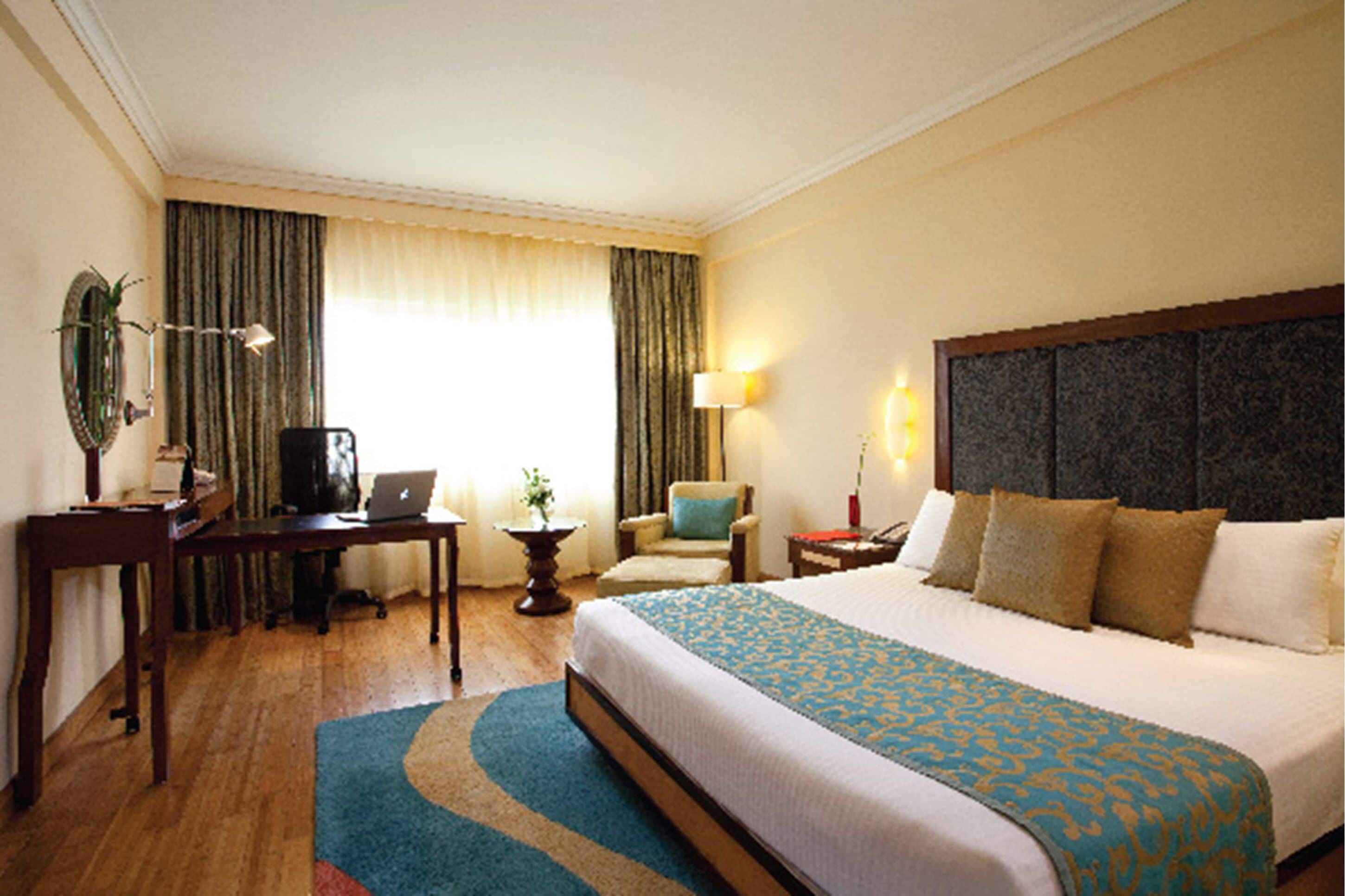 Habitación de hotel con una cama grande, un escritorio con una computadora portátil, una silla, una lámpara y una ventana con cortinas.