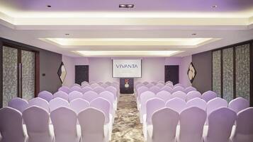 A conference room with rows of white chairs, a projector screen displaying "VIVANTA," and a carpeted floor.