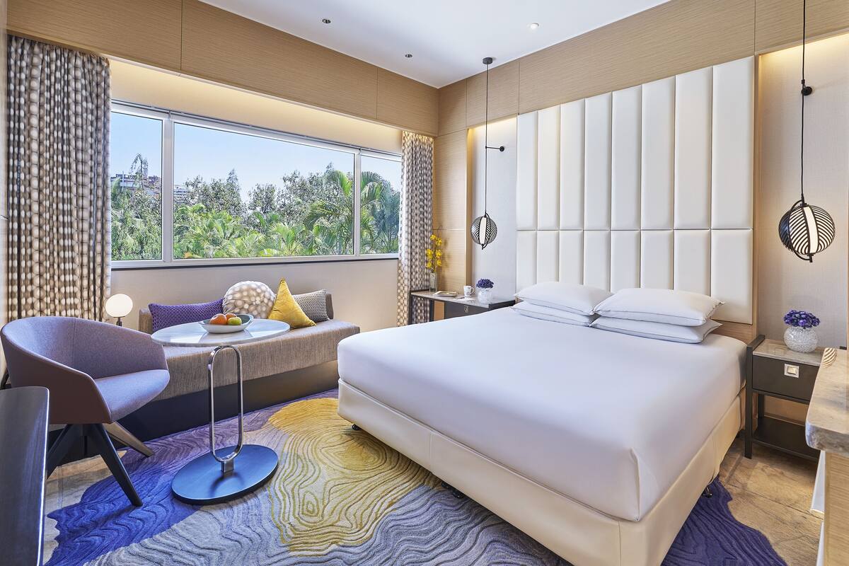 Una habitación de hotel moderna con una cama grande, un área de descanso con una silla y una mesa, y vistas a zonas verdes.