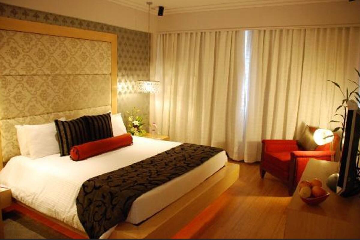 Habitación de hotel con una cama grande, un sillón rojo, una mesita con frutero y una lámpara.