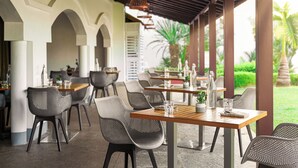 Un espacio exterior cubierto para comer, con mesas y sillas, un techo con luces colgantes y vistas a la vegetación.