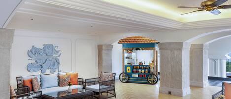 Eine geräumige Lobby mit hoher Decke, Deckenventilator, einer Sitzecke mit Korbmöbeln und einem Imbisswagen.