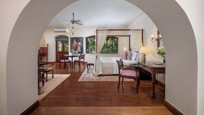 A spacious room with a wooden floor, a bed with a canopy, a dining area with chairs and a table, and a view through an arched doorway.