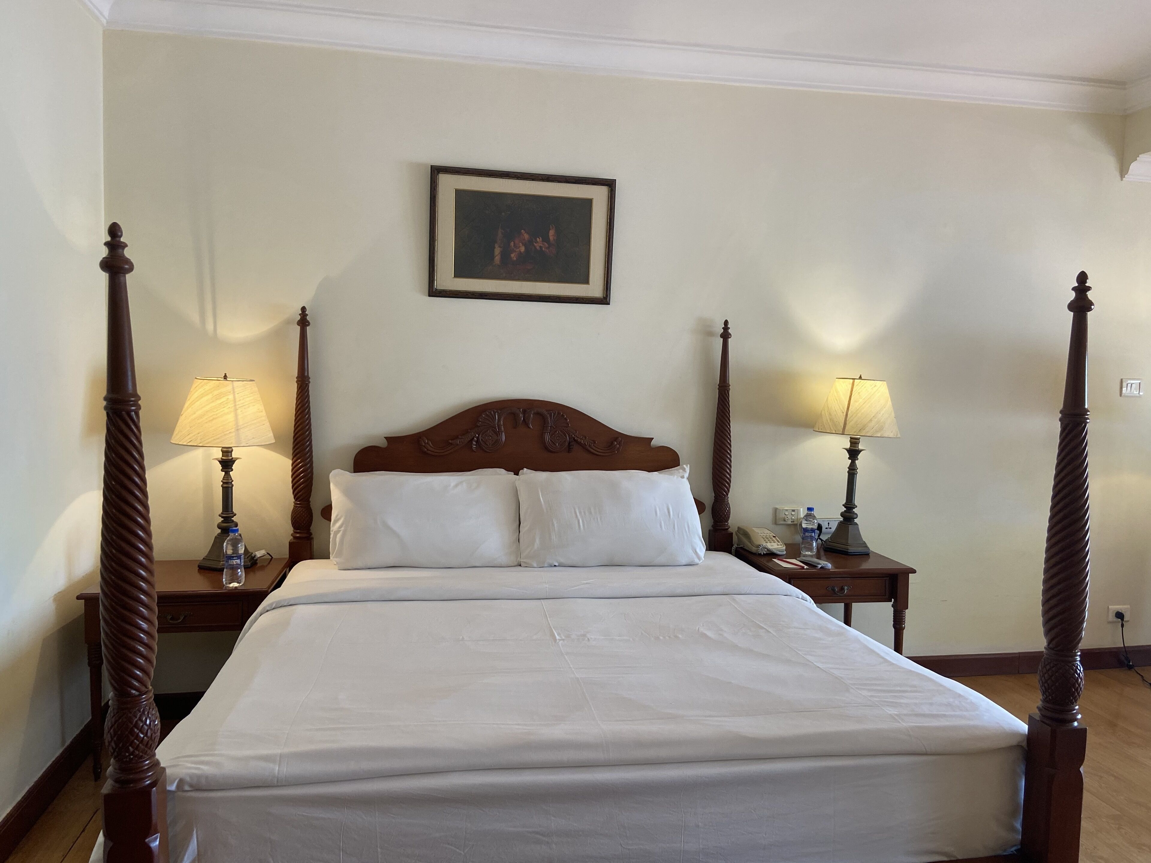Cama bem arrumada com cabeceira de madeira, duas mesas de cabeceira com abajures e um quadro na parede.