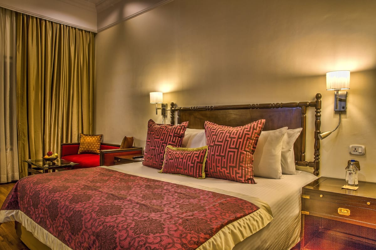A hotel room with a large bed, red and white patterned pillows, a wooden headboard, two bedside lamps, a nightstand, and a red chair.