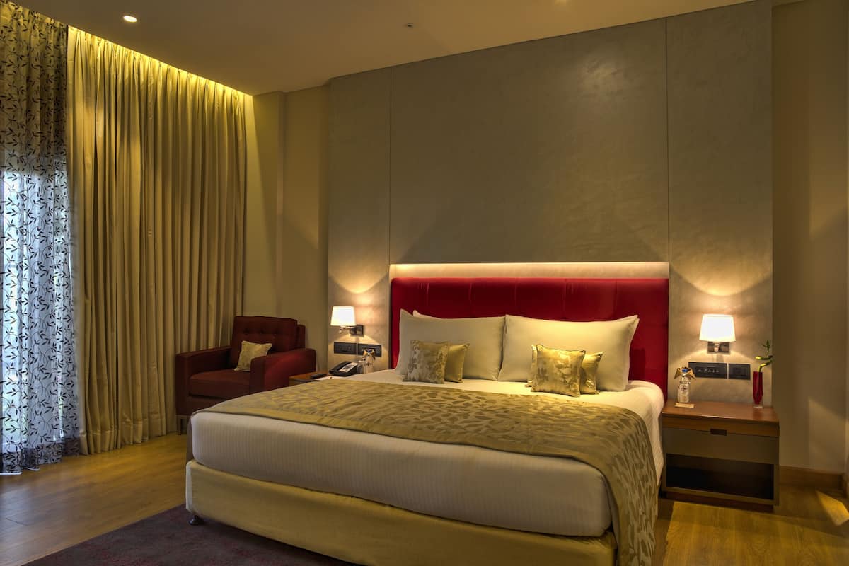 A hotel room with a large bed, a red headboard, two bedside tables with lamps, a red armchair, and a curtain with a leaf pattern.