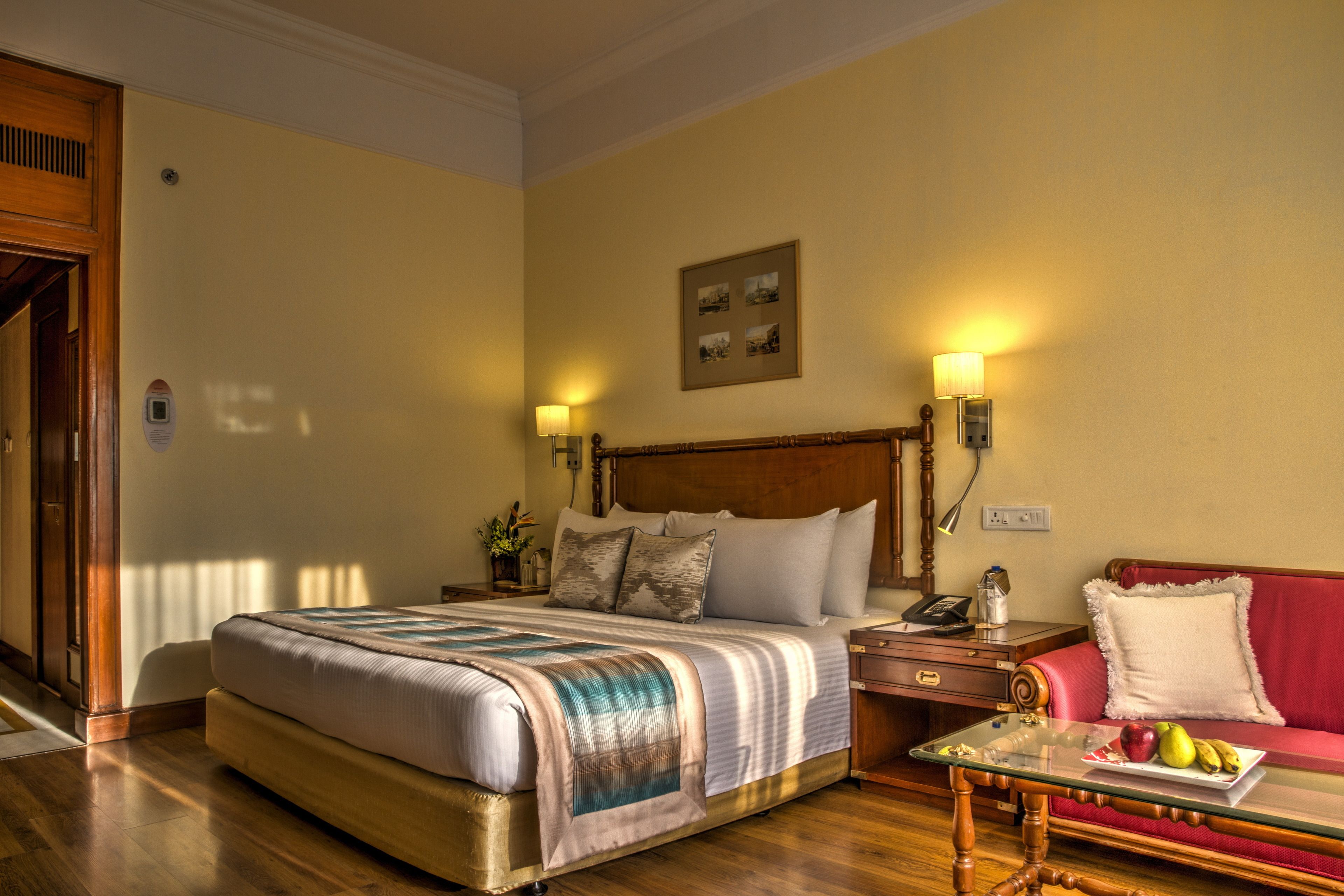 A hotel room with a large bed, a red sofa, a glass coffee table with fruits, and two bedside lamps.