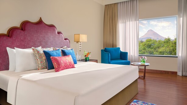 A hotel room with a large bed, a blue armchair, a small table with a lamp, and a window with a mountain view. - Gateway Nashik (Nashik)