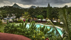Zona de piscina con sillones y sombrillas, rodeada de vegetación exuberante y una montaña al fondo.