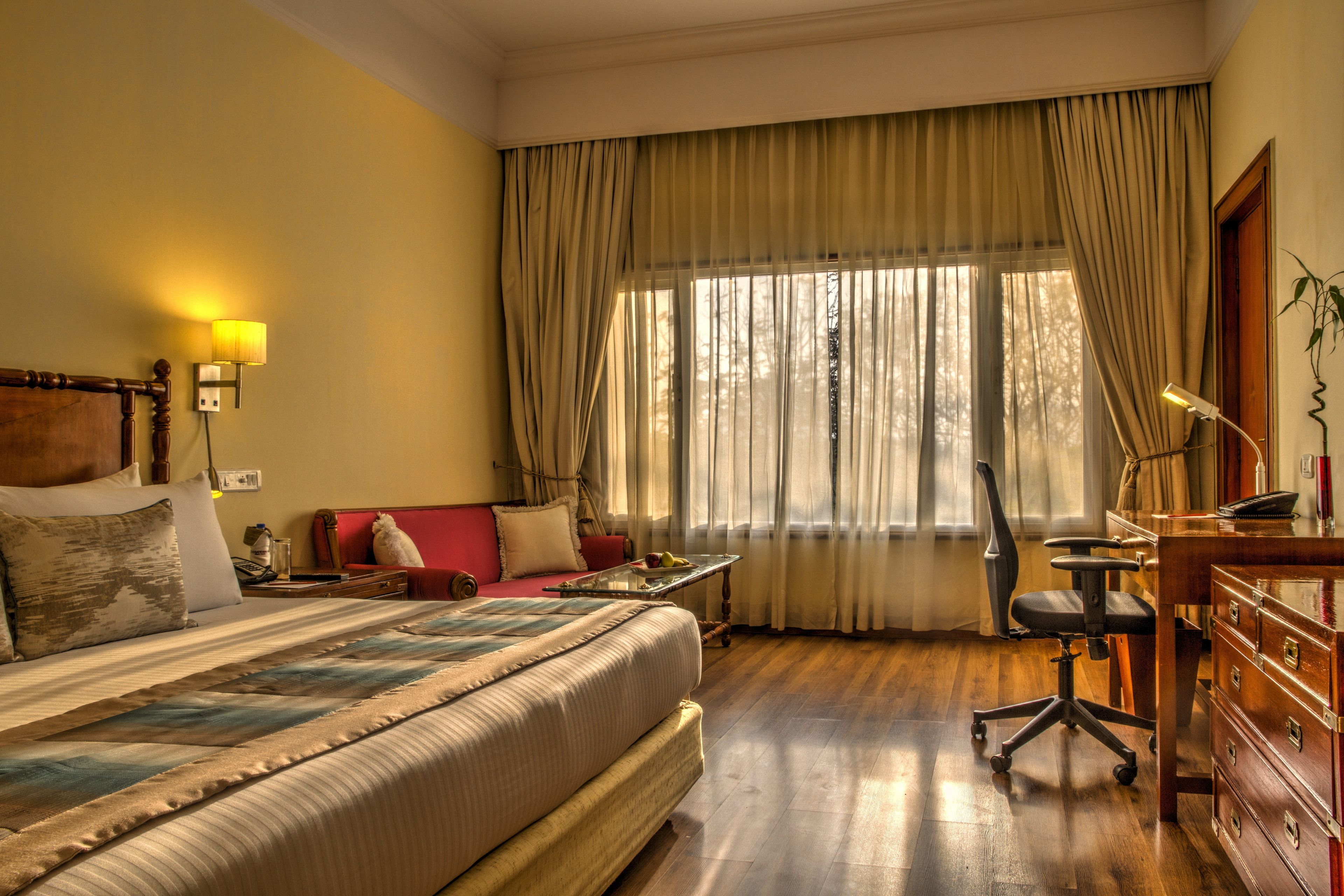 Habitación de hotel con una cama grande, un escritorio con silla, una lámpara y un ventanal con cortinas.