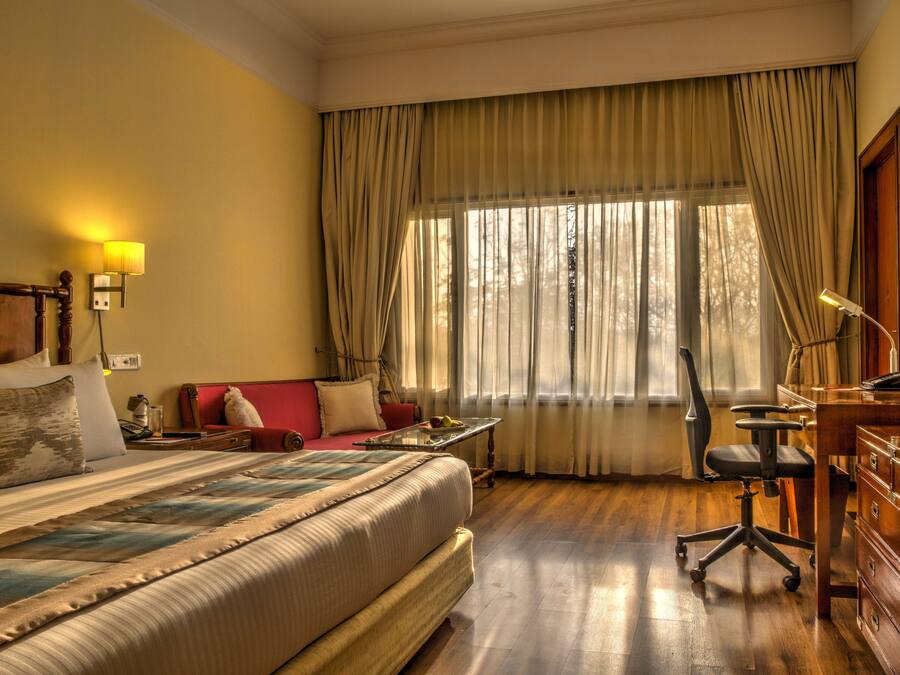 Habitación de hotel con una cama grande, un escritorio con silla, una lámpara y un ventanal con cortinas.