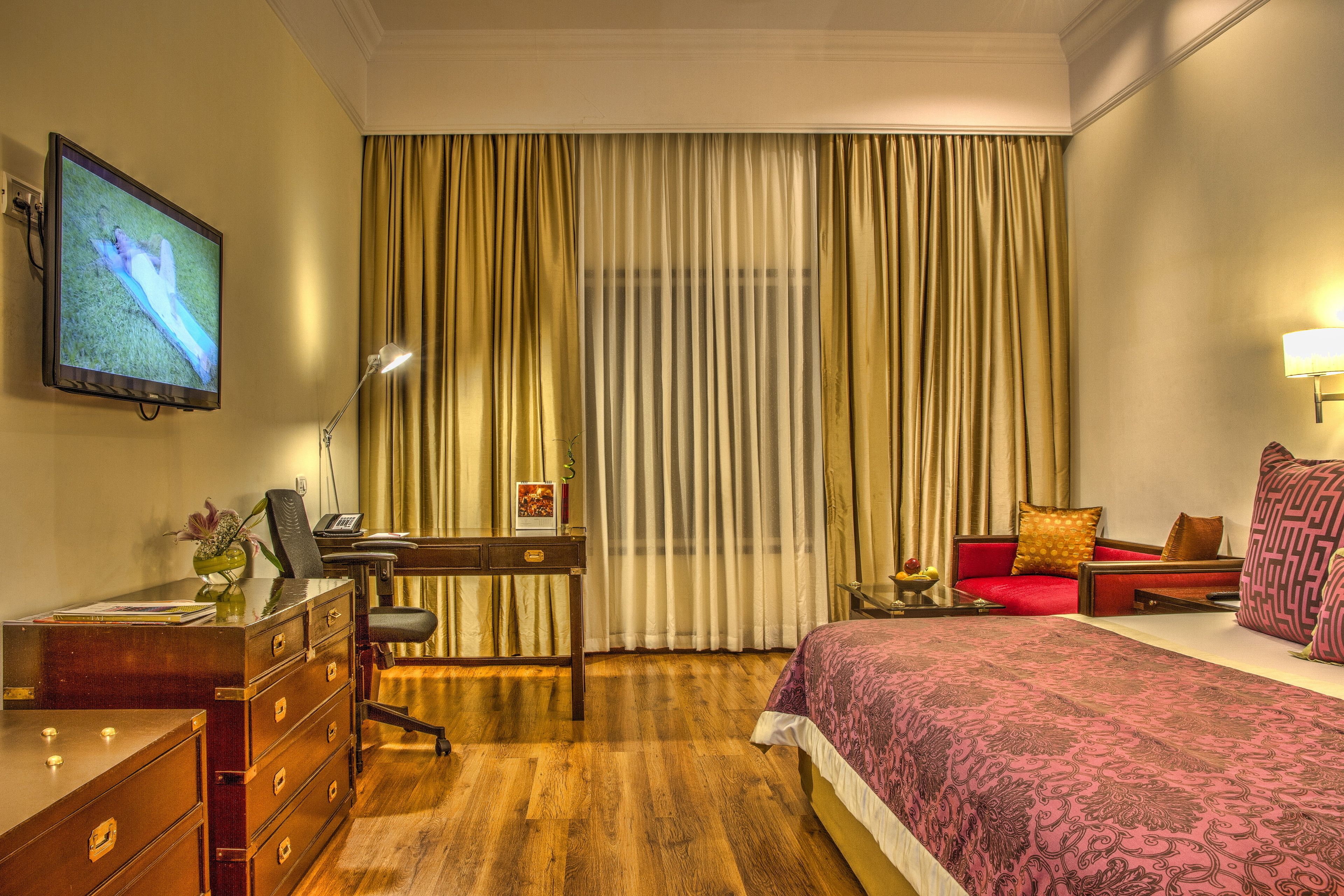 A hotel room with a wooden desk, a chair, a bed with a patterned bedspread, a television on the wall, and curtains.