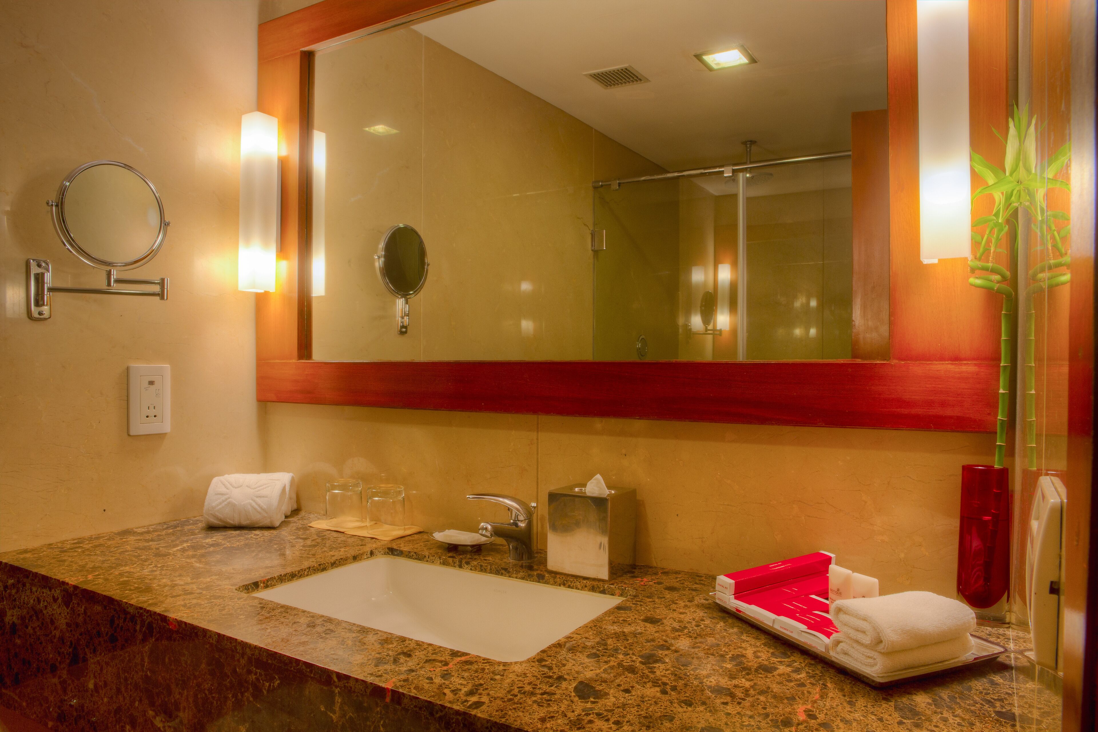 A hotel bathroom with a granite sink, a mirror, a towel rack, a glass of water, and a red vase.
