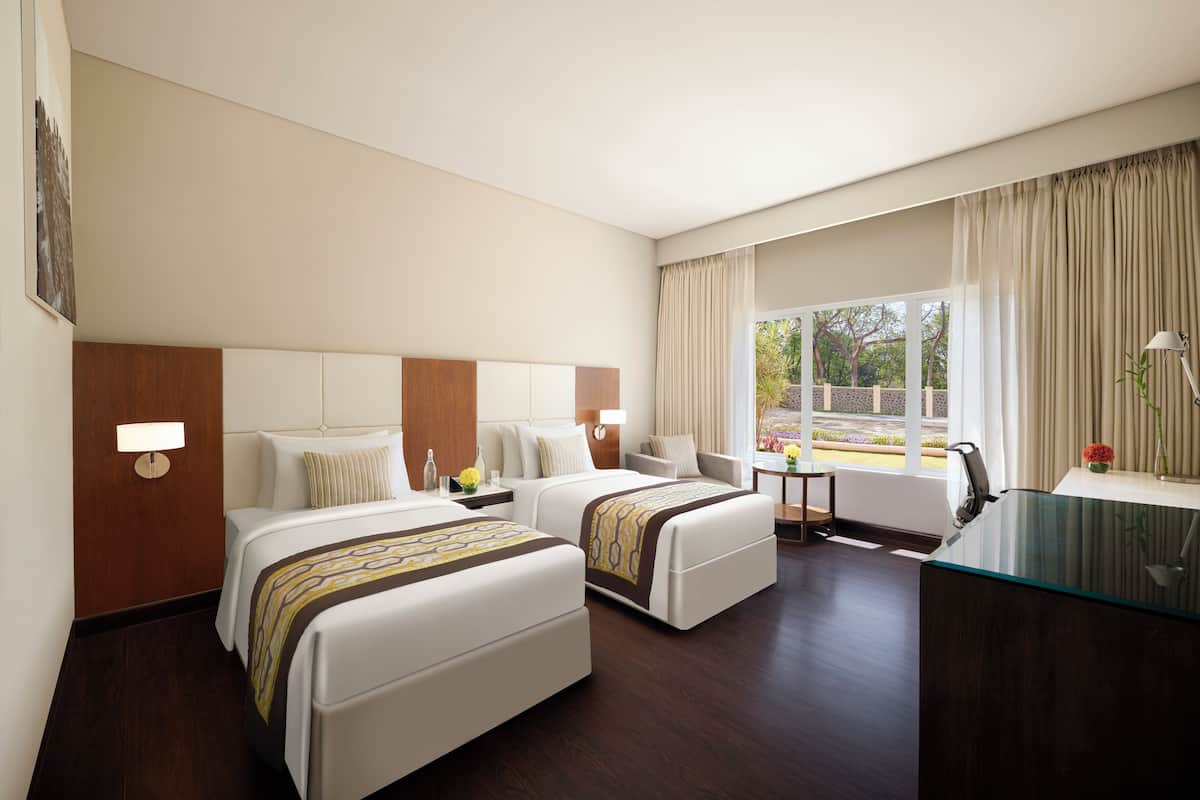 A hotel room with two beds, a desk, a chair, a lamp, and a window with curtains.