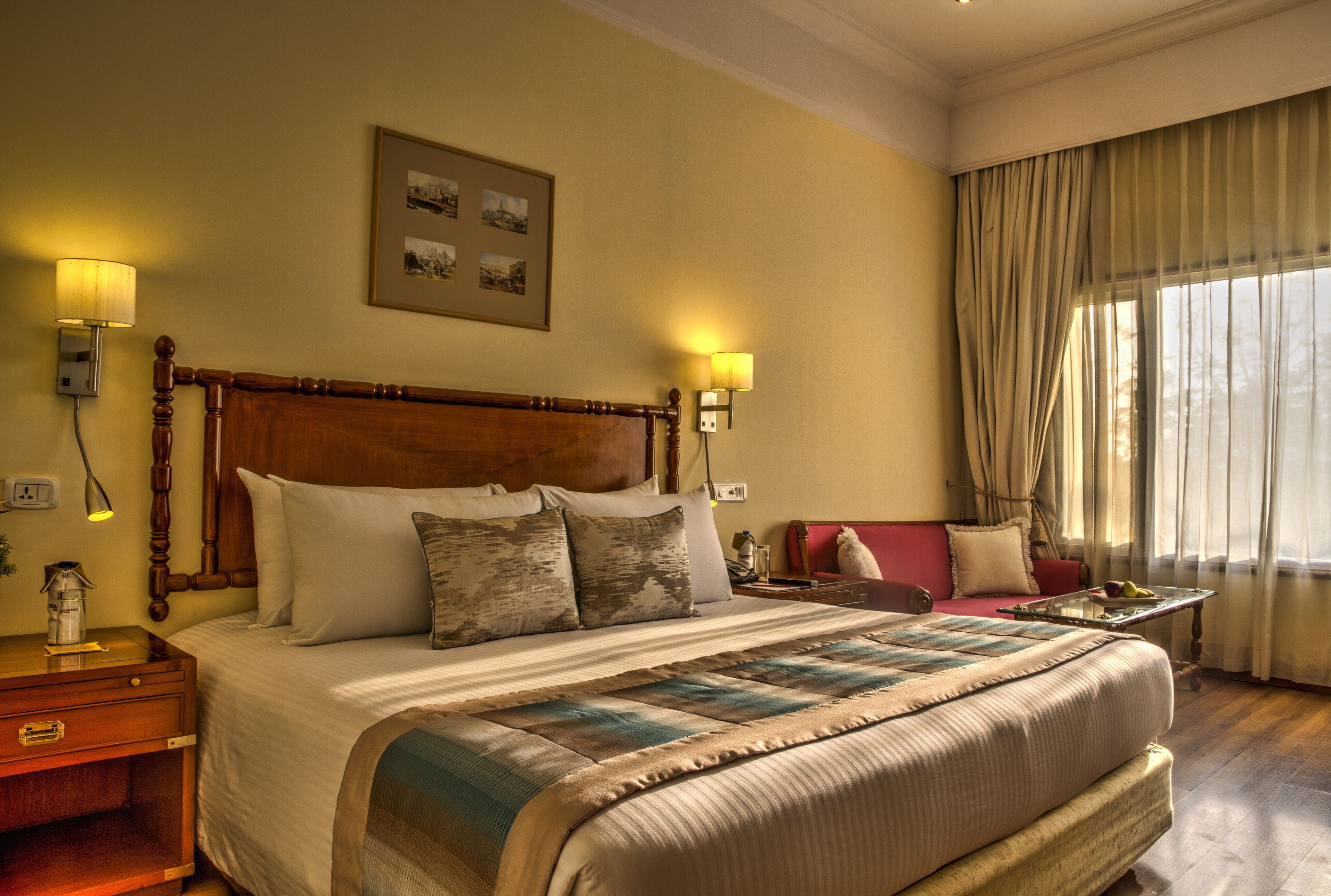 Habitación de hotel con una cama grande, dos lámparas de noche, una mesita de noche, un sofá rojo, una mesa de centro de vidrio y una ventana con cortinas.