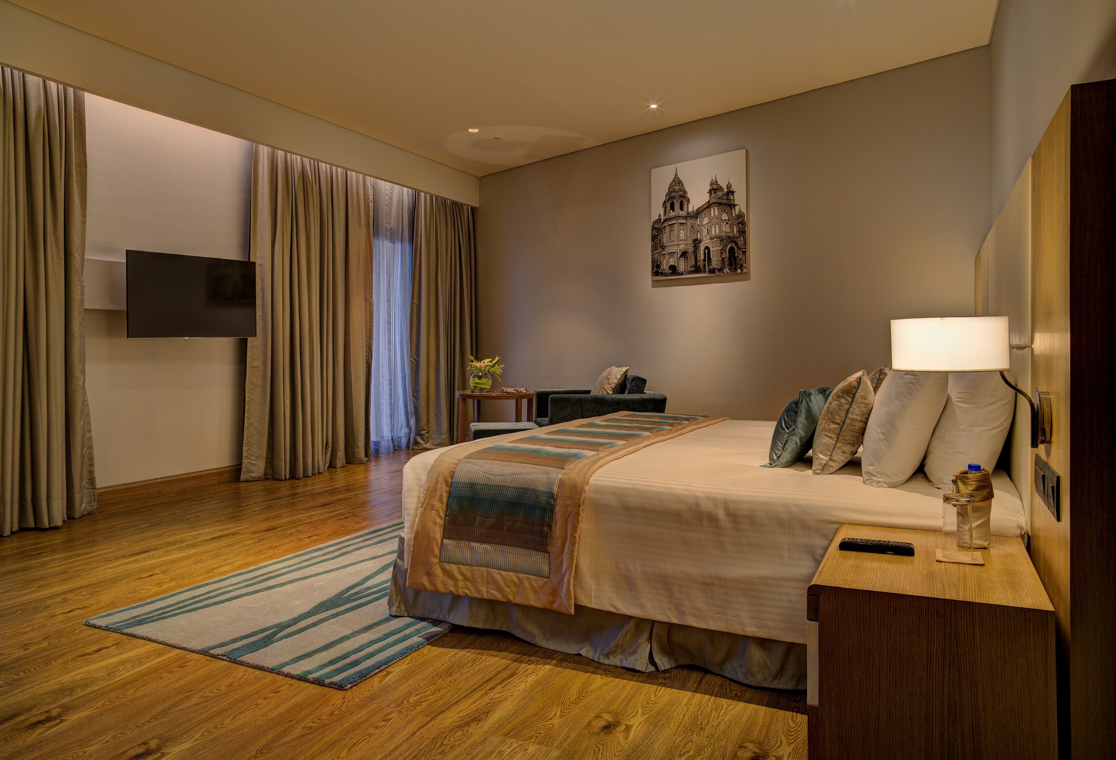 Une chambre d’hôtel avec un lit, un bureau, un téléviseur fixé au mur et un tableau représentant un bâtiment.