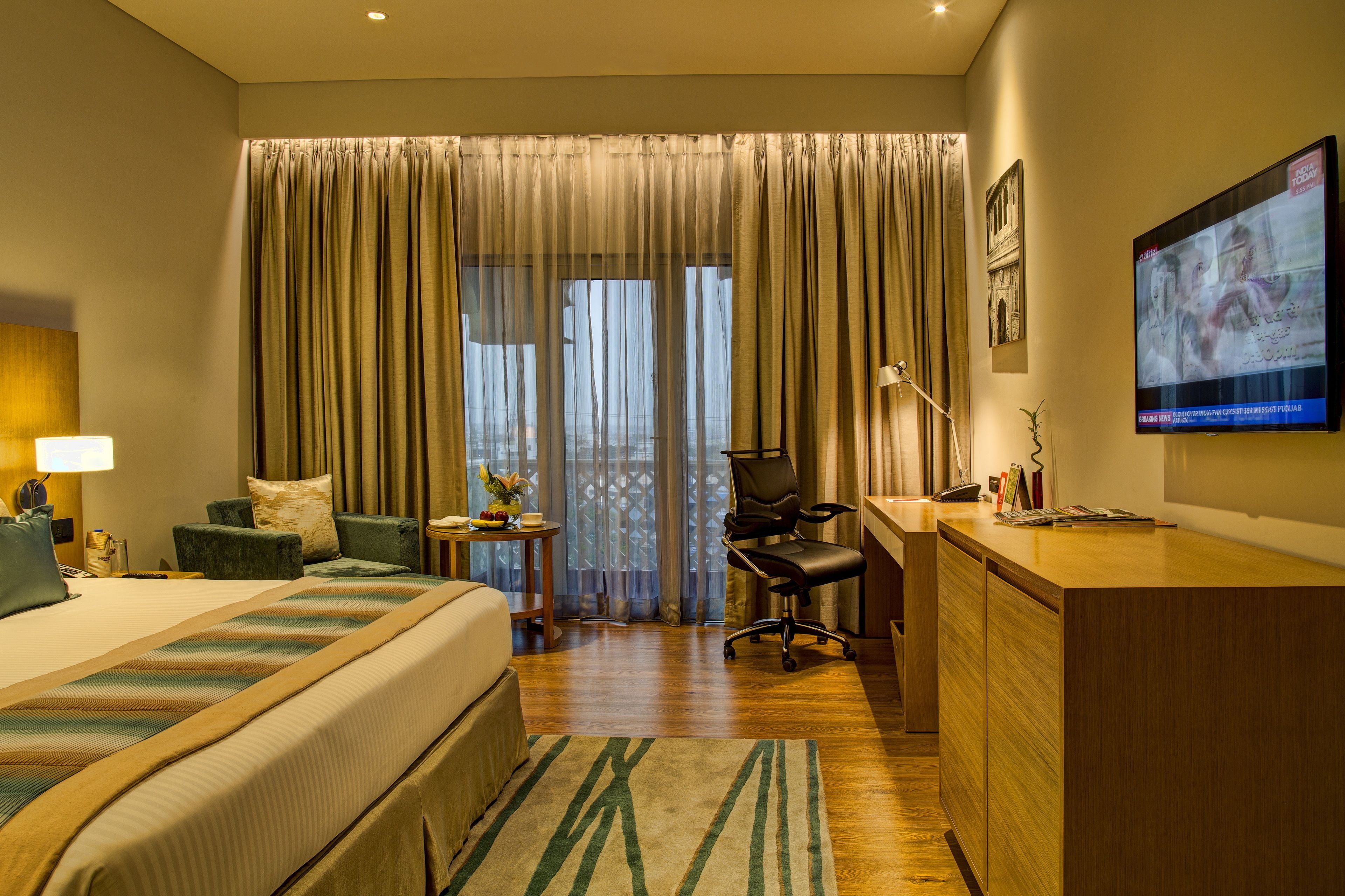 A hotel room with a large bed, a desk, a chair, a TV, and a balcony with curtains.