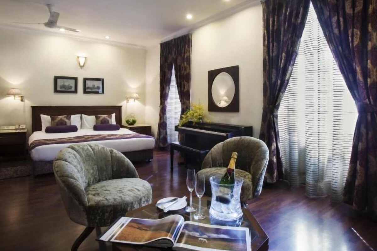 A hotel room with a bed, two armchairs, a small table with a magazine, a bottle of champagne, and a piano.