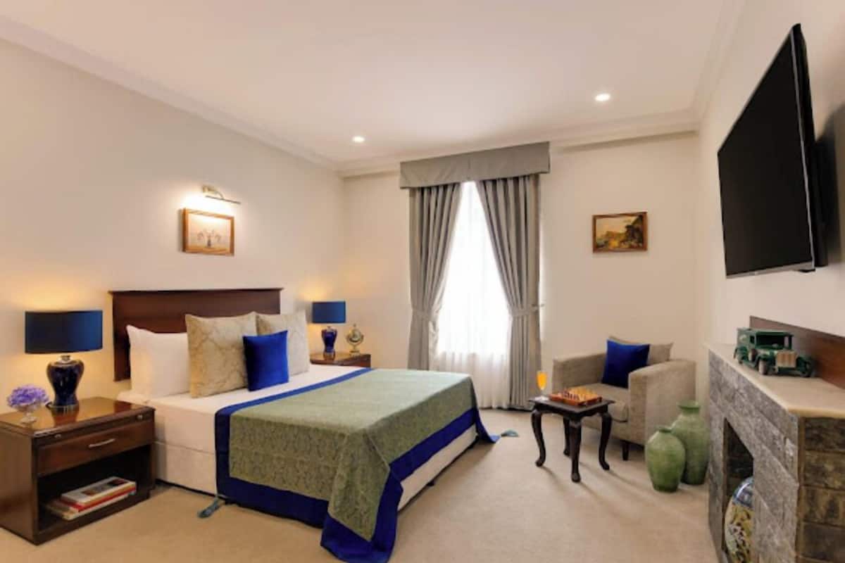 A hotel room with a bed, bedside tables, a TV, a fireplace, and a window with curtains.