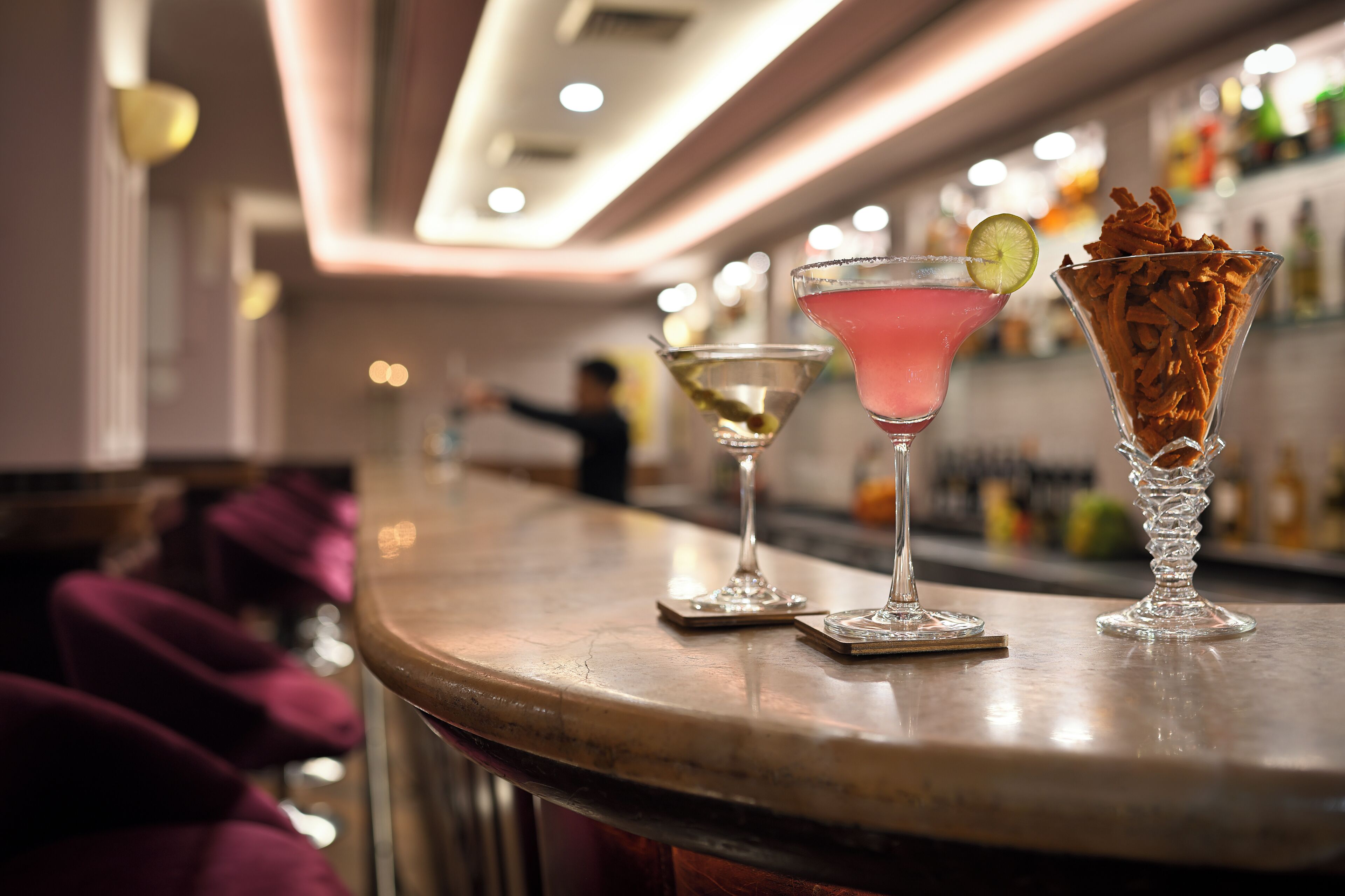 A bar area with three cocktails on the counter, a person in the background, and a bar stocked with bottles.